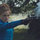 Keine Texte erkennbar. Eine Frau in blauer Kleidung zielt mit einer rauchenden Pistole in freier Natur.