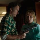 Kein Text erkennbar. Ein Mann in einem karierten Hemd zeigt einer Frau in einem blauen Mantel eine Pistole in einem Wohnmobil.