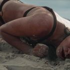 Keine Texte vorhanden. Eine Person liegt mit dem Gesicht im Sand am Strand, trug einen Bikini und wirkt erschöpft.