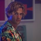 Ein Mann mit welligem blondem Haar trägt ein tropisches Hemd und blickt zur Seite, im Hintergrund bunte Kunstwerke.