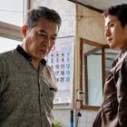 Kalender zeigt Juni und Juli. Zwei Männer stehen in einem schlichten Raum, einer blickt ernst, der andere nachdenklich.