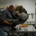 Zwei Männer kämpfen in einer Küche, einer hebt den anderen über einen Tisch. Im Hintergrund Messer und Gemüse.