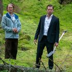 Zwei Männer im Anzug und Outdoor-Kleidung stehen im grünen Wald, einer hält eine Schaufel.