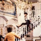 Ein Mann schwingt an einem Kronleuchter über einer Treppe in einem prunkvollen Raum; andere Personen beobachten ihn.