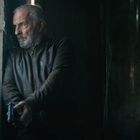 Ein älterer Mann lehnt an einer Wand, hält eine Pistole und schaut ernst aus einem vergitterten Fenster hinaus.