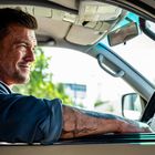 Ein Mann mit Bart sitzt in einem Auto und schaut nach draußen, Arm lässig aus dem Fenster. Tätowierung am Unterarm.