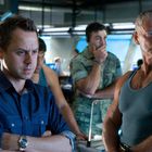 Zwei Männer stehen im Vordergrund: einer in blauer Uniform, der andere im grauen Tanktop. Im Hintergrund sind Soldaten.