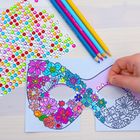 Eine Hand dekoriert eine Blumenmaske mit bunten Glitzersteinen. Daneben liegen farbige Buntstifte.