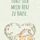 "Bei dir fühlt sich mein Herz zu Hause." Illustration eines Hasen, umgeben von Blumen in Herzform auf pastellgrünem Hintergrund.