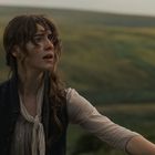 Eine junge Frau mit besorgtem Gesichtsausdruck steht in einer weiten, grünen Landschaft. Sie trägt ein Halstuch und eine Weste.