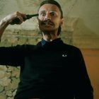 Ein Mann mit Schnurrbart hält einen Revolver an seinen Kopf. Er schaut ernst und trägt einen dunklen Pullover. Im Hintergrund ist eine gemusterte Tapete.