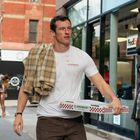 Ein Mann trägt eine Pizzaschachtel von "CLAREMONT PIZZA." Er hat ein kariertes Hemd über der Schulter und geht in einer Stadt.