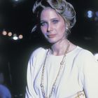 Eine Frau in weißem Outfit mit goldener Kette und passender Handtasche, vor dunklem Hintergrund mit unscharfen Lichtern.