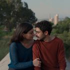 Ein Mann und eine Frau sitzen eng nebeneinander; beide schauen sich liebevoll an. Im Hintergrund sind Bäume sichtbar.
