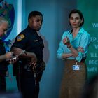 Text: "When 'I' replaced by 'We'... 'Illness' becomes 'Wellness'." Zwei Polizisten mit Notizbuch, Frau mit Namensschild.