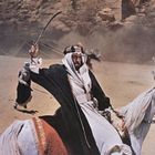 Ein Mann in traditioneller arabischer Kleidung reitet auf einem Pferd und schwingt ein Schwert in einer staubigen Wüstenlandschaft.