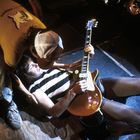 Zwei Personen, eine liegend mit Gitarre, die andere kniet darüber. Der Liegende trägt ein gestreiftes Shirt.