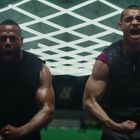 Zwei muskulöse Männer schreien in einem Fitnessstudio. Einer trägt ein Shirt mit Nike-Logo, im Hintergrund sind helle Lichter.