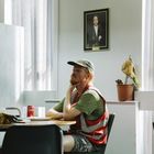 Ein Mann sitzt nachdenklich an einem Tisch mit einer Cola-Dose in einem Raum. Porträt einer Frau an der Wand.
