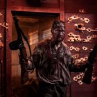 Ein Mann mit zwei Gewehren, Schlamm bedeckt, steht vor einer rot beleuchteten Wand voller Einschusslöcher.