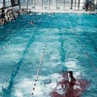 Eine Gruppe von Menschen schwimmt in einem großen Hallenbad. Eine Person steht im Zentrum, umgeben von rotem Wasser.