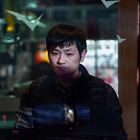 Ein Mann steht hinter einer Glasscheibe in dunkler Umgebung, trägt eine Jacke und wirkt nachdenklich.