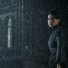 Eine Frau steht im Regen vor einem dunklen, historischen Gebäude, schaut zur Seite.
