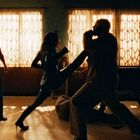 Personen im Schatten liefern sich einen Kampf vor großen, hellen Fenstern. Eine Person tritt die andere.