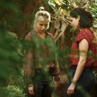 Zwei Frauen stehen in einem dichten, grünen Wald und scheinen in ein Gespräch vertieft zu sein.