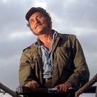 Ein Mann mit Kappe und grünlicher Jacke steht auf einem Boot und schaut nach oben. Seile verlaufen parallel im Vordergrund.