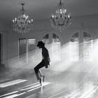 Ein Tänzer mit Hut posiert in einem hell erleuchteten Raum mit Kronleuchtern und Fenstern, durch die Lichtstrahlen fallen.