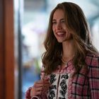 Ich kann keine erkennbare Schrift im Bild finden. Eine Frau mit langen Haaren lächelt, trägt ein kariertes Jackett und Perlenkette.