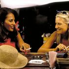 Zwei Frauen sitzen lächelnd an einer Bar, mit Gläsern und einem Strohhut, beide halten eine Zigarette.