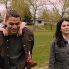 Ein Mann trägt ein Kind auf dem Rücken, neben ihm geht eine Frau auf einer grünen Wiese, im Hintergrund Bäume und Häuser.