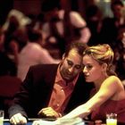 Zwei Personen, eng aneinander gelehnt, an einem Casinotisch mit Getränken und Spielchips. Im Hintergrund weitere Personen.