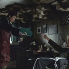 Ein Mann in rotem Overall zeigt einem im Sessel sitzenden Mann einen grünen Ofenhandschuh. Die Atmosphäre wirkt angespannt.