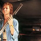 Eine Frau in einem blauen Hemd hält ein langes Gewehr. Sie trägt eine gelbe Uhr und wirkt konzentriert.