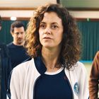 Ein Frau in heller Jacke vor Tennishalle, flankiert von drei Männern im Hintergrund. Alle blicken ernst.