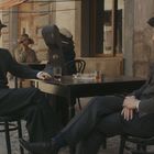 Zwei Männer in Anzügen sitzen draußen an einem Tisch auf einer Kopfsteinpflasterstraße.