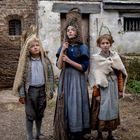 Drei Kinder in rustikaler Kleidung stehen vor einem alten Steingebäude, eines hält einen Besen.