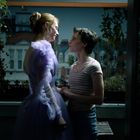 Zwei Personen stehen auf einem Balkon, liebevoll einander zugewandt. Eine trägt ein fliederfarbenes Kleid, die andere ein gestreiftes Shirt.