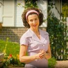 Frau mit rosa Stirnband und Kleid steht lächelnd vor einem weißen Lattenzaun mit Blumen und einem Haus im Hintergrund.