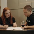 Eine Frau und ein Mann sitzen an einem Tisch mit Dokumenten. Die Frau zeigt mit einem Stift auf das Papier. Ein Glas Rotwein steht daneben.