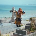 Eine Person in einem eleganten Kleid steht auf einem Friedhof mit Meerblick, ausdrucksvoll posierend.
