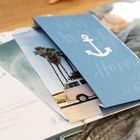 Ein blauer Umschlag zeigt einen weißen Anker und den Text "Happiness is". Darunter ein Foto mit Palmen und einem pinken Bus.