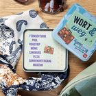 "WORT & weg: Das verrückte Wortspiel für unterwegs" steht auf einer Blechdose. Daneben ein geöffneter Deckel mit Wörtern.
