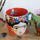Ein Becher mit buntem Frauenporträt und Blumenkranz, daneben ein grüner Becher mit Rehmotiv und Pinsel im Hintergrund.