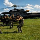 Text: "OM-HAT", "AIRBUS". Vier Soldaten in Tarnkleidung vor einem Helikopter auf einem Feld, bereit zum Einsatz.