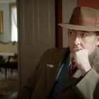 Es gibt keine erkennbaren Texte im Bild. Ein Mann mit Hut sitzt in einem altmodischen Raum, im Hintergrund Möbel und Gemälde.