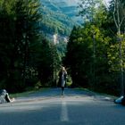 Eine Frau geht auf einer Waldstraße. Links und rechts liegen zwei Personen auf dem Boden. Hintergrund: Berge und Bäume.